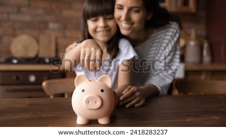 Saving money. Happy hispanic mother hug preteen daughter dropping coin into piggybank support child in wish to make savings teach economy financial planning. Soft focus on cute piggy bank toy on table Royalty-Free Stock Photo #2418283237