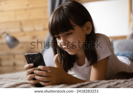 Child and cellphone. Smart little hispanic girl relax lie on bed look on cell screen watch video cartoon. Small gen z kid addicted to gadgets spend leisure time using phone having fun playing web game
