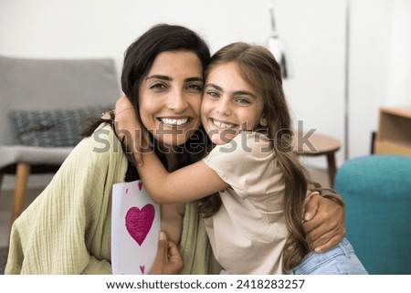 Cheerful Latin mom and excited daughter girl holding greeting card with heart and hugging, looking at camera with toothy smiles. Happy embracing mum due to mothers day, posing for family portrait