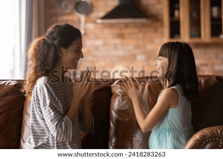 Clap your hands. Joyful preteen girl play patty cake with caring mexican nanny babysitter sit on sofa at country cottage. Loving stepmother entertain little foster child having fun enjoy simple game Royalty-Free Stock Photo #2418283263