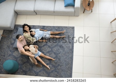 Happy middle aged Latin couple of parents and two kids resting on carpeted warm floor, looking up at camera, smiling, laughing, enjoying home family leisure. Top view, full length shot