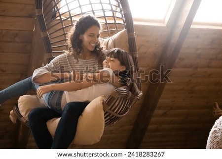 Affectionate moment. Loving hispanic female foster mom swing in cozy wicker chair hold small adopted kid daughter on knees tickle laugh having fun. Happy older younger sisters play indoors. Copy space Royalty-Free Stock Photo #2418283267