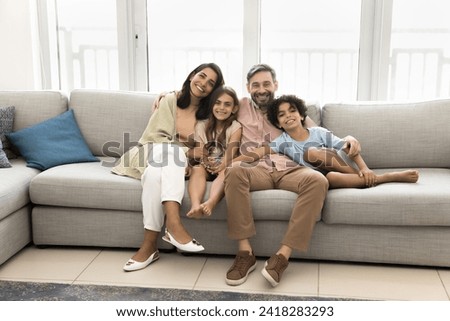 Cheerful young parents and preteen children resting on home couch, looking at camera, smiling, enjoying being in comfortable new apartment, promoting mortgage, affordable property buying for family