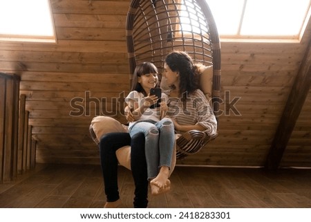 Smiling single latina mother relax in rattan chair swing at comfy attic hold preteen daughter on lap use smartphone. Friendly mom and child girl talk discuss news at social network play cellphone game
