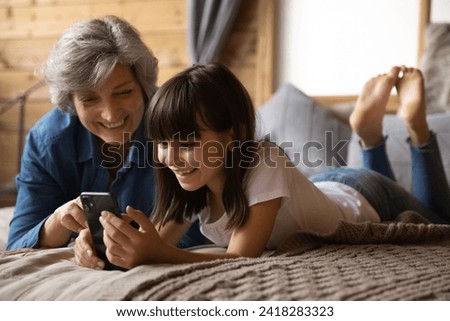 Happy preteen kid granddaughter rest on large bed with loving retired hispanic granny buying toys on ecommerce web site play cellphone game in online app. Mature babysitter use phone with little girl
