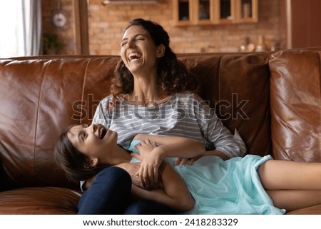 Tickle and giggle. Happy family of latina foster mom and cute girl adopted child having fun relax on couch at home laugh enjoy funny active game. Excited small kid cuddle play on sofa with adult nanny Royalty-Free Stock Photo #2418283329