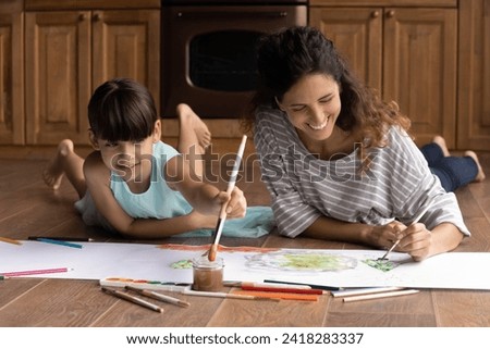 Quiet activity. Inspired hispanic woman babysitter engaged in drawing colorful picture with little preteen age girl. Happy adult elder sister lie on floor with heating help younger kid to paint poster