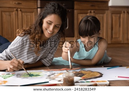 Funny artists. Smiling latina family young foster mother and small daughter adopted child paint big picture with watercolors. Young mexican nanny play on warm wooden floor with little girl draw sketch