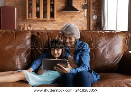 Aged hispanic female babysitter spend time on sofa with preteen girl using tablet pc device watch cartoon play game. Mature granny and little grandchild learn foreign language in online app for kids