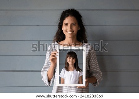 When I was a child. Headshot portrait of pretty hispanic woman posing with her childhood photo. Smiling young latina mother stand by grey wall look at camera show picture of preteen little daughter