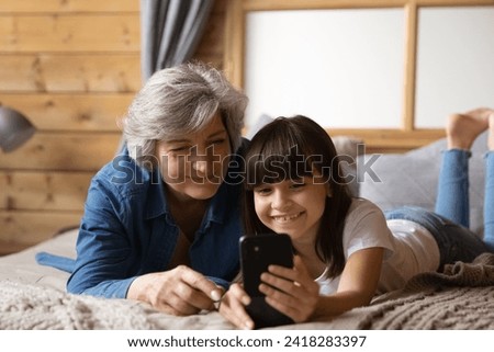 Happy little latin girl lie on bed close to old age grandmother show new photo video at social network account. Joyful preteen grandchild make cute selfie on cell webcam together with senior granny