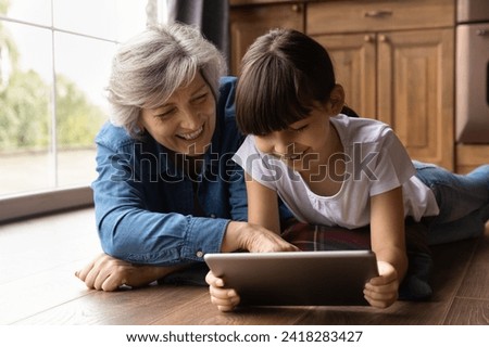 Elderly latina grandmother rest on warm floor with preteen grandkid touch tablet screen in girl hands explain learning material in playful way. Little girl play online game with loving granny on pad