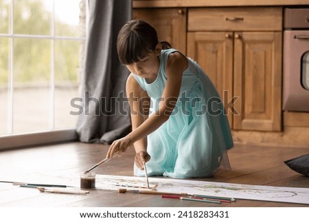 Happy kid. Preteen hispanic girl engaged in creative activity draw funny picture with brush watercolors in large light kitchen with floor heating. Little child play quiet game enjoy painting at home