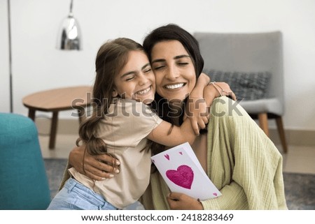 Happy excited mommy and cheerful child girl celebrating mothers day, birthday, 8 march, holding handmade paper greeting card with drawn purple heart, hugging with cheek touch and closed eyes