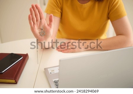 A woman with a laptop holds a religious Jewish symbol while sitting at a table in a home kitchen. An adult female businesswoman works from home, a remote office Royalty-Free Stock Photo #2418294069