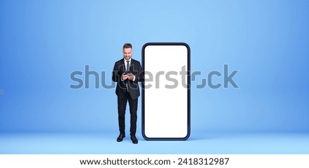 Smiling businessman in formal suit near big mockup copy space phone screen, blue background. Concept of business network, financial and internet communication Royalty-Free Stock Photo #2418312987