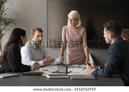 Senior business group leader woman standing at meeting table, listening to speaking younger employees, holding brainstorming meeting. Diverse team sharing ideas for cooperation Royalty-Free Stock Photo #2418316959