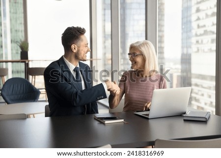 Positive confident elder and younger colleagues discussing collaboration, partnership, laughing, giving handshake. Cheerful business partners shaking hands at workplace, getting agreement