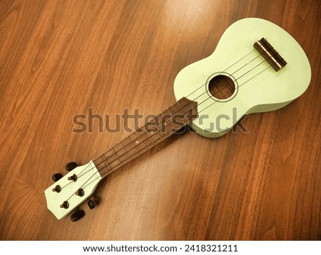 A pastel green ukulele with a wooden fretboard rests on a wooden floor, its strings inviting a musical touch. Royalty-Free Stock Photo #2418321211