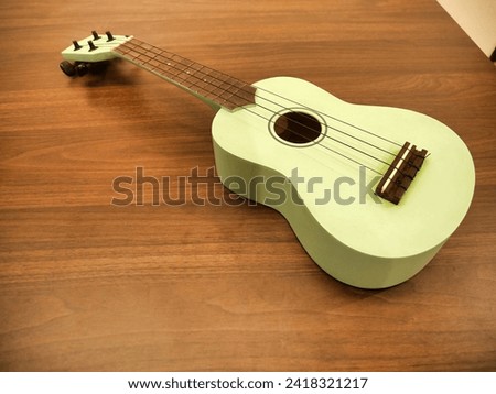 A pastel green ukulele with a wooden fretboard rests on a wooden floor, its strings inviting a musical touch. Royalty-Free Stock Photo #2418321217