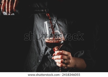 woman hands pouring red wine into a glass from a bottle on black background Royalty-Free Stock Photo #2418346055