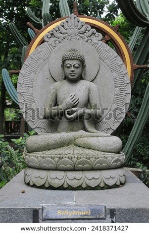 A stone carved statue of the Buddha or Siddhartha Gautama meditating in a garden background. Photo taken from the front at an eye level angle