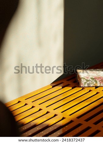 Sunny product display: Yellow bench against a concrete wall with an intricately illustrated flowery box. Cozy ambiance with raindrop-kissed window shadows. Ideal for showcasing your products. Royalty-Free Stock Photo #2418372437