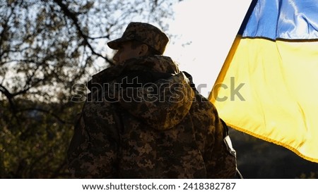 Young male military in uniform walks with waving flag of Ukraine at countryside. Soldier of ukrainian army going with blue-yellow banner outdoor. Concept of victory against russian aggression.