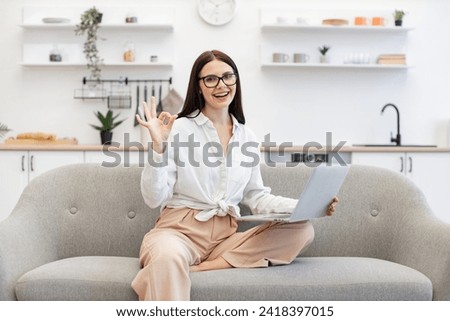 Happy brunette businesswoman sitting on comfy couch and using portable laptop showing sign ok. Excited caucasian female feeling inspired with new idea during freelancing at cozy home.