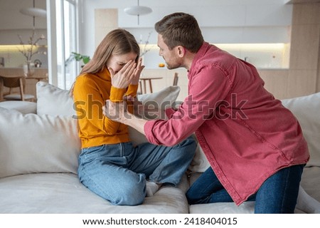 Aggressive stressed tyrant man husband shouts grabs wife by arms. Unhappy scared woman crying closing face by hands sitting at home. Domestic physical, emotional violence, family quarrel, gaslighting. Royalty-Free Stock Photo #2418404015
