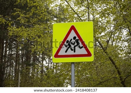Road sign Denoting Be careful, children, on pole, that is installed near schools. Yellow square, red triangle, white, with the image of running people. Safety, Traffic warning, autumn forest.