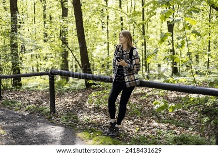 Young happy woman with smartphone wears a black backpack walking in the forest. Spring time. 30s Women in sport clothes hiking in forest. Wanderlust travel concept, atmospheric moment