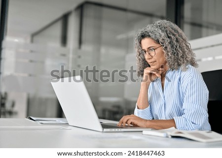 Busy mature business woman working in office using laptop. Middle aged old professional lady executive manager entrepreneur looking at computer thinking on digital technology strategy sitting at desk. Royalty-Free Stock Photo #2418447463