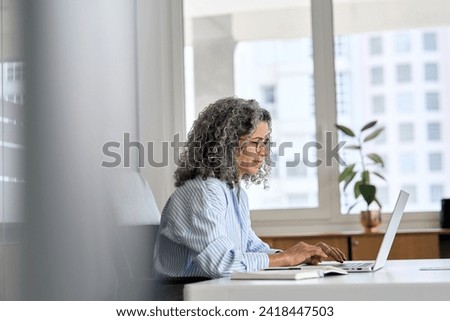 Busy mature senior business woman working in office using laptop. Middle aged old professional lady executive manager looking at computer digital technology sitting at desk. Authentic candid shot. Royalty-Free Stock Photo #2418447503