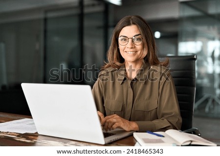Happy smiling mature middle aged professional business woman investor manager executive or lawyer attorney adviser looking at camera at workplace working on laptop computer in office, portrait. Royalty-Free Stock Photo #2418465343