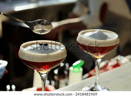 Espresso martini, pictured with selective focus, pictured at a craft cocktail bar. The beverage is served in a coupe glass and topped with frothy foam and espresso coffee beans for garnish.