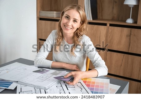Smiling young female freelancer holding color swatches at home workspace. Drawings, architecture plans, pantone palette on table. Creative ideas for interior design at house