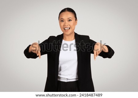 Enthusiastic glad millennial Asian businesswoman with a broad smile pointing downwards with both hands, highlighting something below, against a neutral grey background, studio