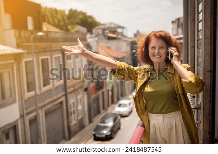 Senior woman standing on sunny balcony, talking on cellphone and waving hand, with city streets in the background, happy elderly lady feeling joyous and connected, greeting friend, copy space Royalty-Free Stock Photo #2418487545