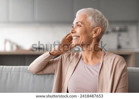 Portrait of happy european mature lady looking away with smile and touching face, enjoying her time and relaxation at home indoor, free space. Woman with gray short hair posing for photo indoor Royalty-Free Stock Photo #2418487653