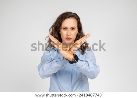 A determined confident caucasian young woman in a blue shirt makes an X sign with her arms, signaling denial, rejection, or a boundary being set, with a serious expression