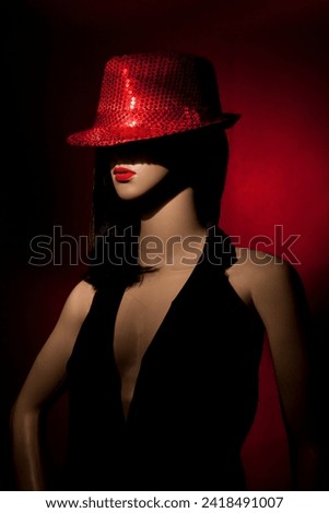Plastic woman mannequin with clack long hair, red lipstick, and wearing a bright red sequins hat posing on a red background in a one light studio setting Royalty-Free Stock Photo #2418491007