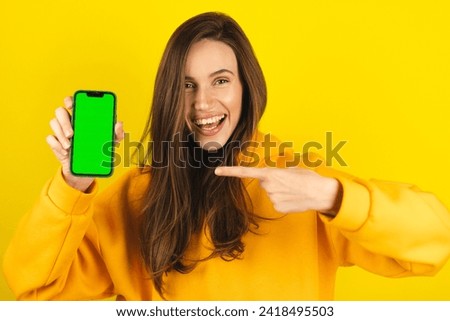 Young developer woman hold smartphone with green screen chroma key mock up recommend good application promotional sale offer, point to phone screen. Brunette girl isolated on yellow studio background.