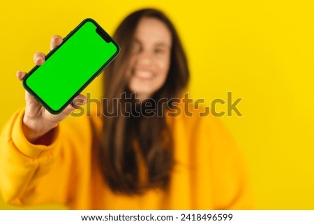 Excited young woman in yellow hoody hold and show blank phone green screen with copy space for app, isolated on yellow background. Happy amazed woman with wow emotions, recommend app, focus on phone.