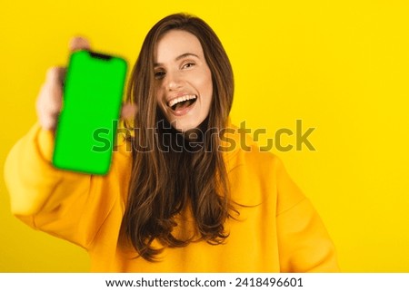 Excited young woman in yellow hoody hold and show blank phone green screen with copy space for app, isolated on yellow background. Happy amazed woman with wow emotions, recommend app or some product.