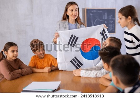 Joyful young female teacher showing flag of South Korea to schoolchildren preteens during history lesson in classroom Royalty-Free Stock Photo #2418518913