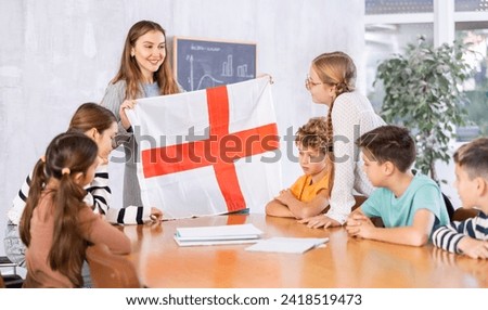 Young female teacher showing flag of England to schoolchildren preteens during history lesson in classroom Royalty-Free Stock Photo #2418519473