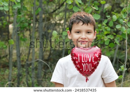 Cute smiling boy in white t-shirt is holding a red heart shaped balloon for St. Valentine's day with words "I love you" standing in the park with green trees.