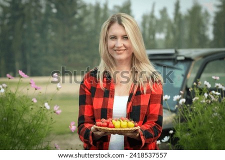 A female farmer is holding a plate with assorted fresh homegrown red and yellow pear shaped cherry tomatoes in the garden, blooming wildflowers and a vehicle in the background. Royalty-Free Stock Photo #2418537557