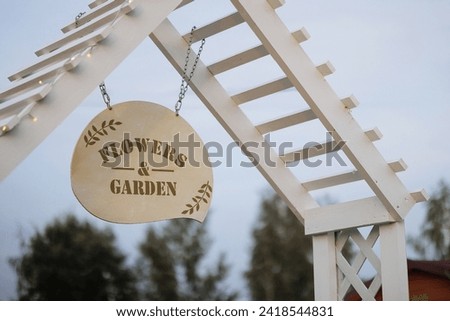 A round Flowers and Garden sign hangs from a white, wooden structure. Concept for garden beautification and outdoor events.